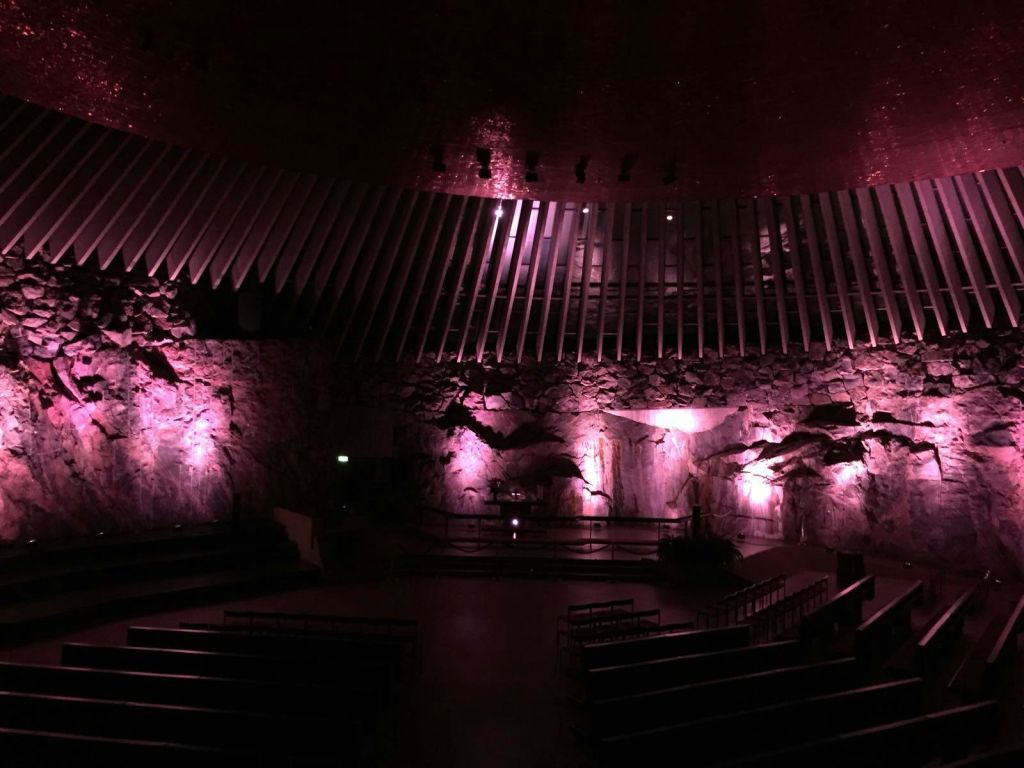 암벽, 보라색 조명, 층층이 배치된 좌석이 있는 조명이 희미한 강당입니다. 스테이지 위의 커다란 갈비뼈 모양의 빔 곡선.