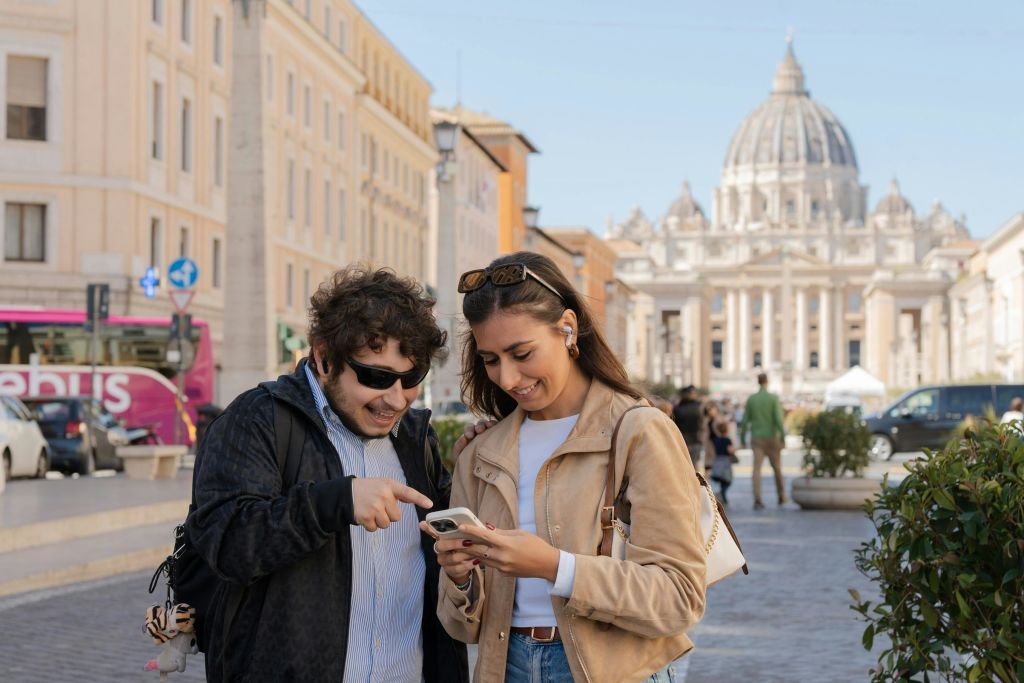 선글라스를 착용한 남성과 이어폰을 착용한 여성이 도시의 거리에 서서 함께 미소를 짓고 스마트폰을 바라보고 있습니다.
