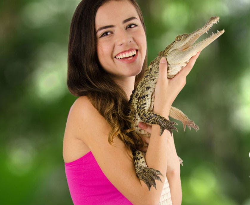 분홍색 상의를 입은 웃는 여성이 흐릿한 녹색 배경에 작은 악어를 들고 있습니다.