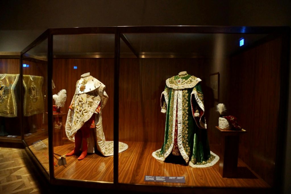 빨간색과 흰색, 녹색과 흰색 두 개의 화려한 머리 없는 왕실 가운이 나무 바닥의 유리 케이스 안에 전시되어 있습니다.