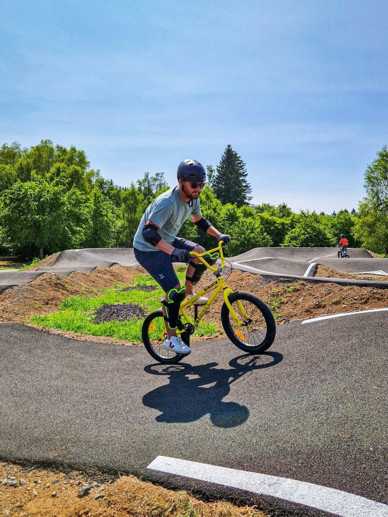 햇살이 내리쬐는 공원의 펌프 트랙에서 노란색 BMX 자전거를 타는 사람이 헬멧과 보호 장비를 착용하고 있습니다.