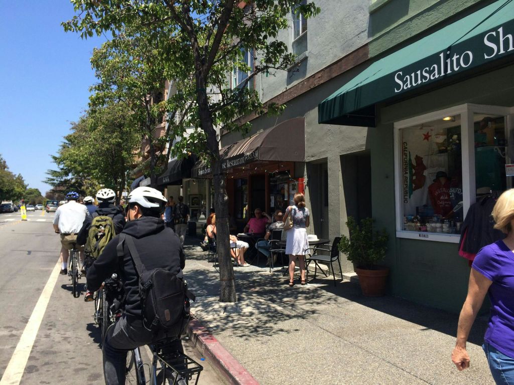 상점과 카페가 있는 거리를 따라 자전거를 타는 사람들 ('소살리토' 표지판이 있는 거리 포함) 과 근처의 보행자.