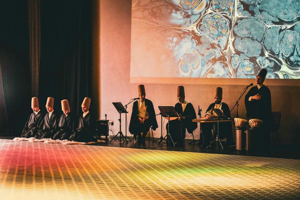 전통 복장을 한 뮤지션들이 무대에서 다채로운 추상적 프로젝션 아래 악기를 들고 공연을 펼칩니다.