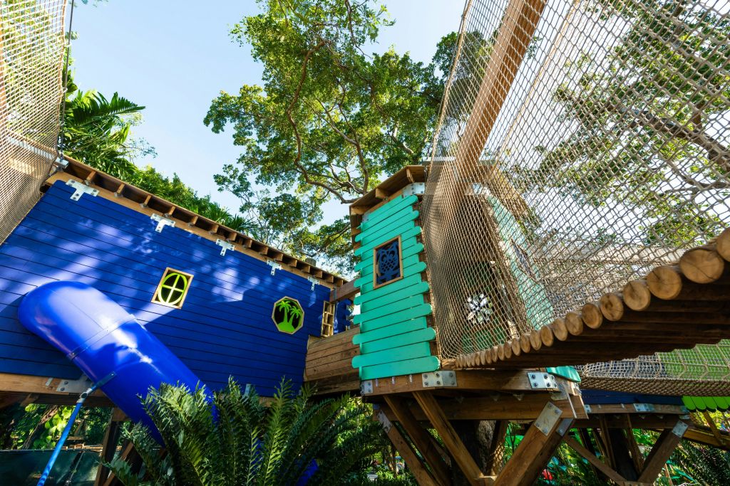 울창한 녹음과 나무로 둘러싸인 로프 브리지가 있는 파란색과 청록색 트리하우스가 특징인 나무 놀이 구조.