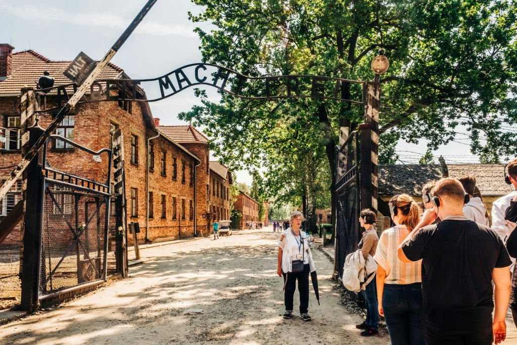 햇살이 내리쬐는 야외 환경에서 '아르비트 마흐트 프레이 (Arbeit Macht Frei)' 라는 문구가 있는 역사적인 출입문 근처의 한 무리의 사람들.