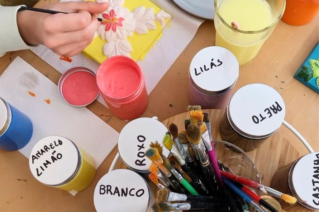 노란 캔버스에 꽃을 그리는 손. "LILÁS", "ROXO", "BRANCO" 라고 표시된 페인트병과 다양한 브러시가 보입니다.