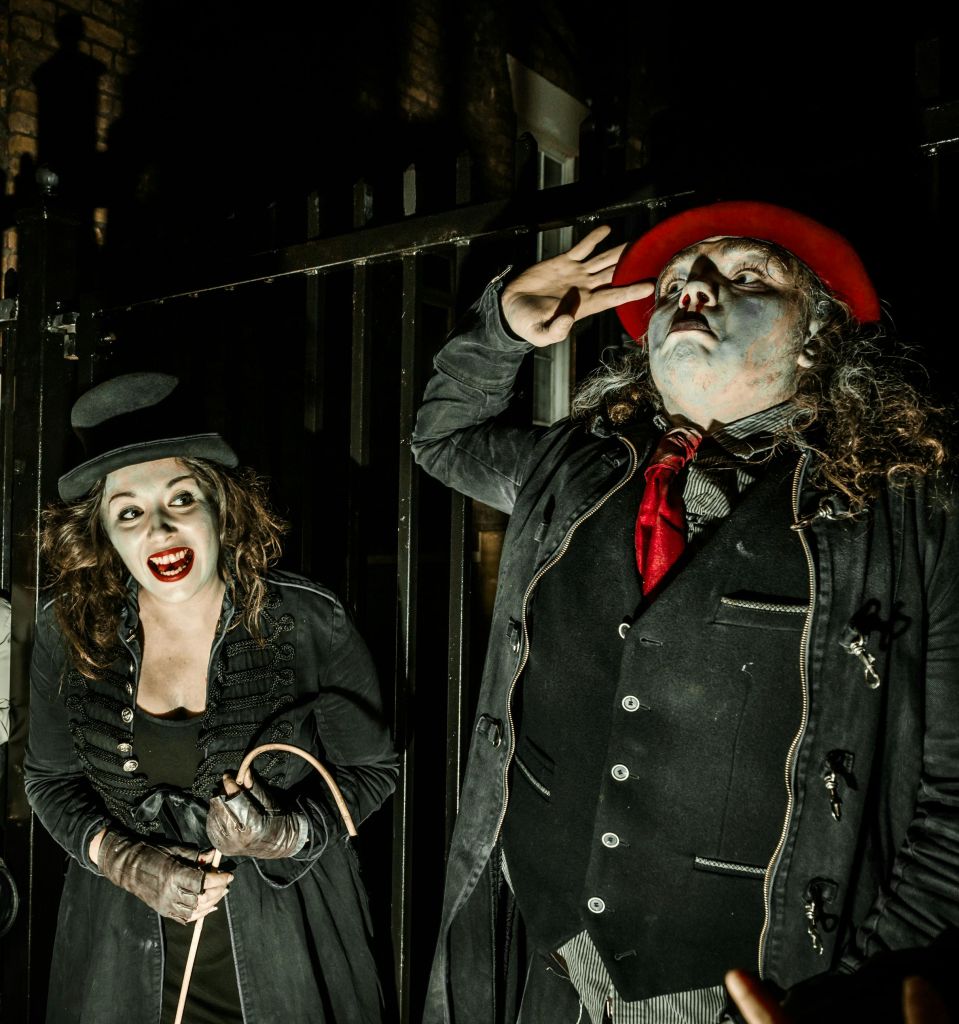 빨간 모자를 쓴 연극 메이크업과 의상을 입은 두 사람이 어둡고 막힌 배경을 배경으로 나란히 서 있습니다.