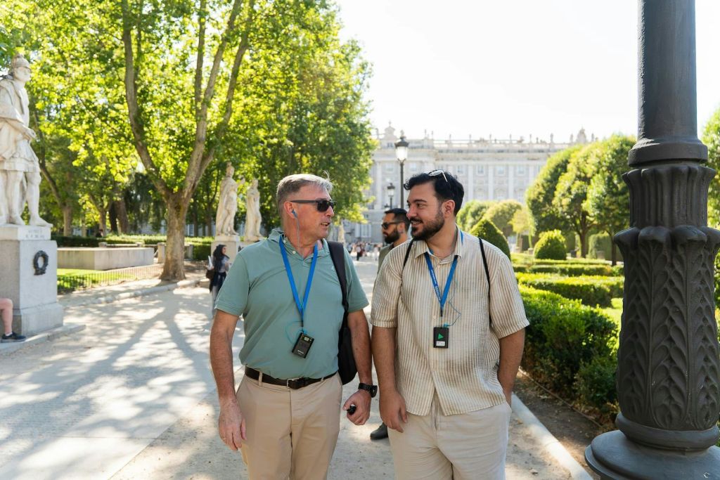 커다란 건물을 배경으로 깔끔한 경관을 자랑하는 공원에서 두 남자가 산책하며 이야기를 나누고 있습니다. 둘 다 투어 가이드 장치를 착용합니다.
