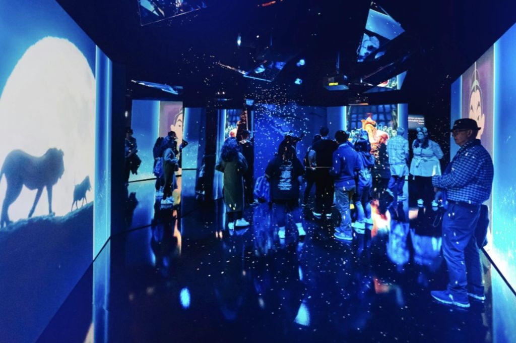 파란색 조명이 있는 어두운 방에 서 있는 한 무리의 사람들이 이미지와 비주얼이 표시된 화면으로 둘러싸여 있습니다.