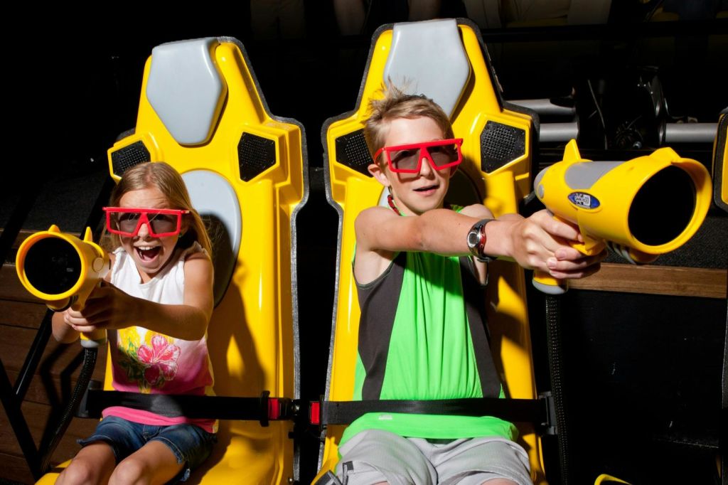 빨간색 3D 안경을 쓰고 노란색 게임용 의자에 앉아 열정적으로 장난감 총을 겨냥하는 두 어린이.