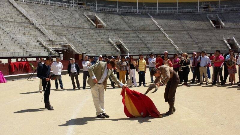전경의 두 사람이 빨간색과 노란색 망토를 들고 투우 시위를 벌이고 있으며, 텅 빈 경기장에서 군중들이 지켜보고 있습니다.