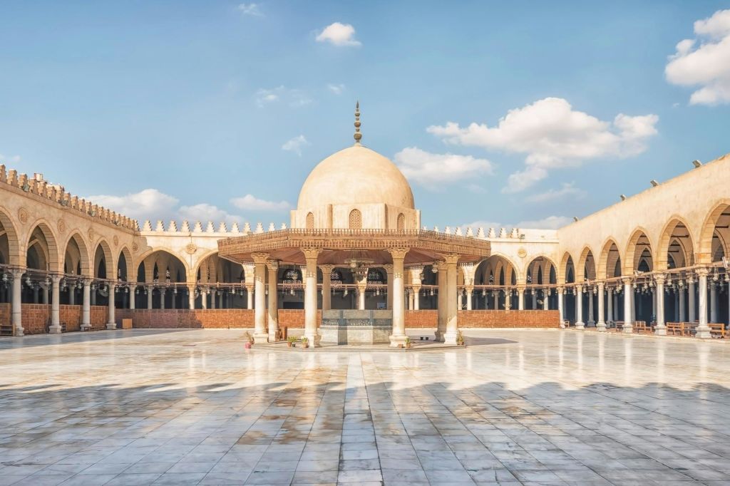 구름 몇 개가 있는 푸른 하늘 아래, 아치와 기둥으로 둘러싸인 중앙 돔 구조의 모스크 마당.