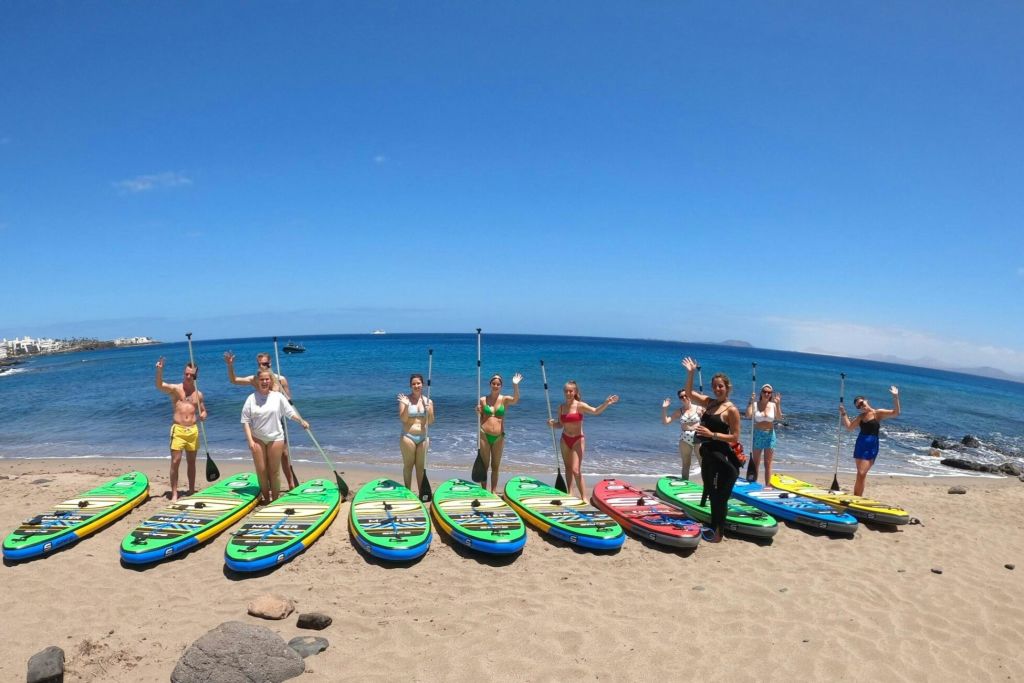 한 무리의 사람들이 패들보드를 들고 모래 해변에 서 있습니다. 패들보드를 앞에 두고 바다를 배경으로 서 있습니다.