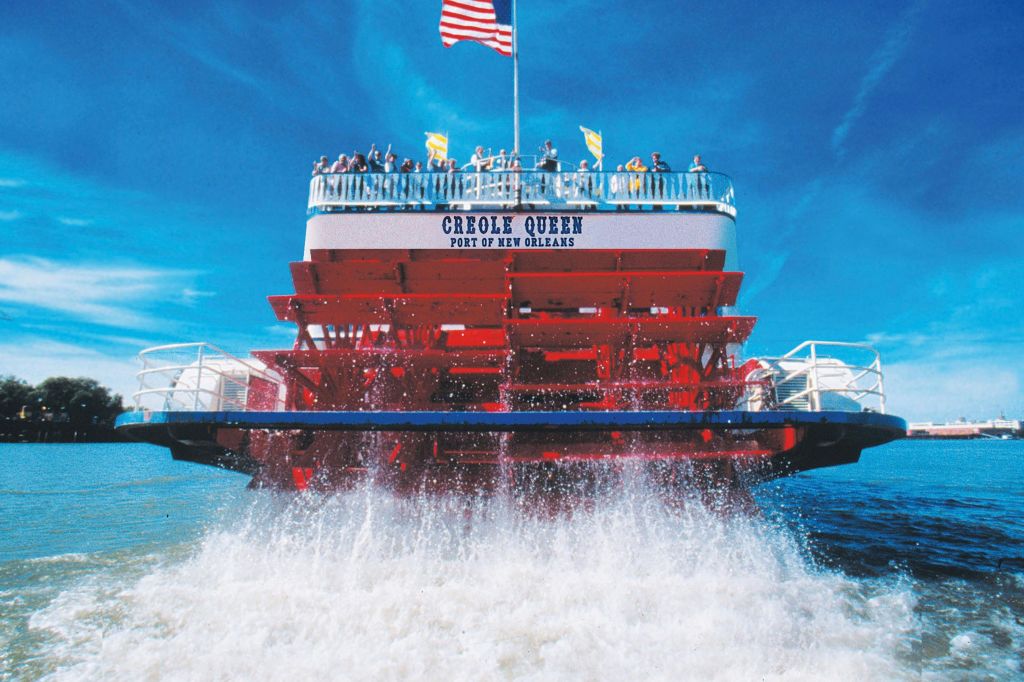 위쪽 갑판에는 사람들이 있고 위쪽에는 맑고 푸른 하늘을 배경으로 미국 국기가 있는 크리올 퀸 강 보트의 빨간 패들휠이 있습니다.