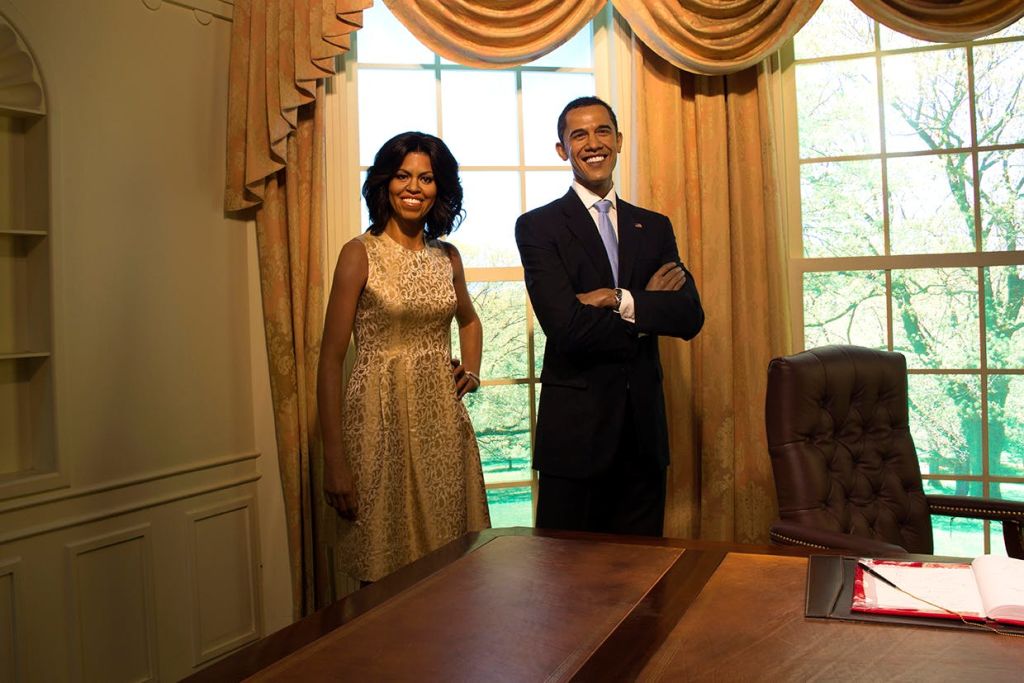 양복을 입은 남성과 베이지색 드레스를 입은 여성이 커다란 창문과 커튼을 배경으로 책상 근처에 서서 미소를 짓고 있습니다.