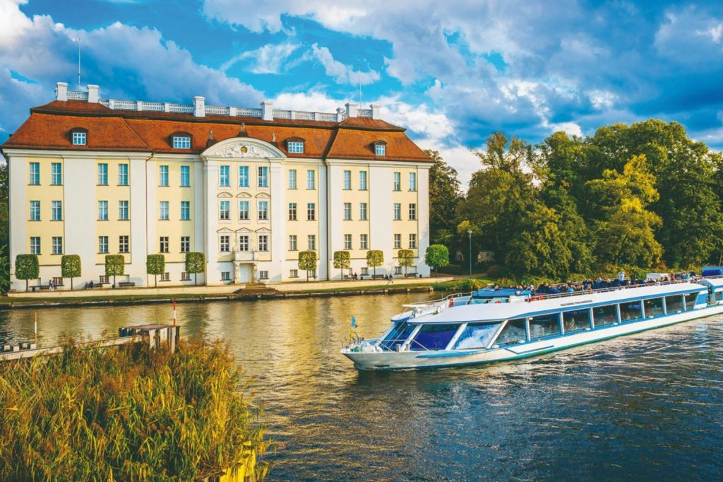 보트는 부분적으로 흐린 하늘 아래 나무와 녹지로 둘러싸인 붉은 지붕의 크고 유서 깊은 건물을 지나 강을 따라 이동합니다.