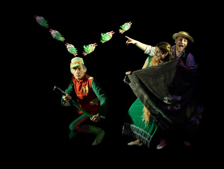 화려한 의상을 입은 세 명의 배우가 어두운 배경을 배경으로 공연하며, 한 명은 포인팅을 하고 다른 한 명은 소품과 천을 들고 있습니다.