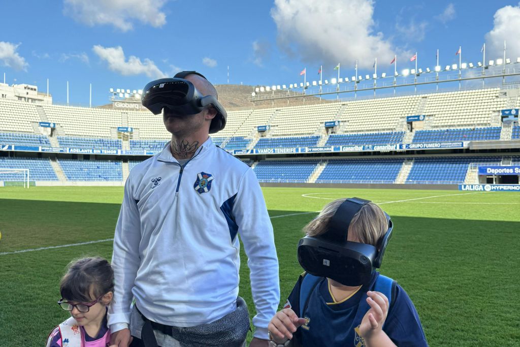 축구장에는 VR 헤드셋을 착용한 세 사람이 서 있으며, 경기장과 푸른 하늘이 배경으로 펼쳐집니다.