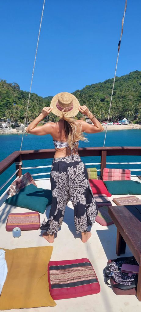 여름 복장을 한 여성이 모자를 쓰고 화려한 쿠션이 있는 보트 갑판에 서서 열대 섬과 해변을 내려다보고 있습니다.