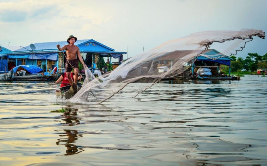작은 배에 선 한 남성이 집과 배를 배경으로 어망을 물에 던집니다.
