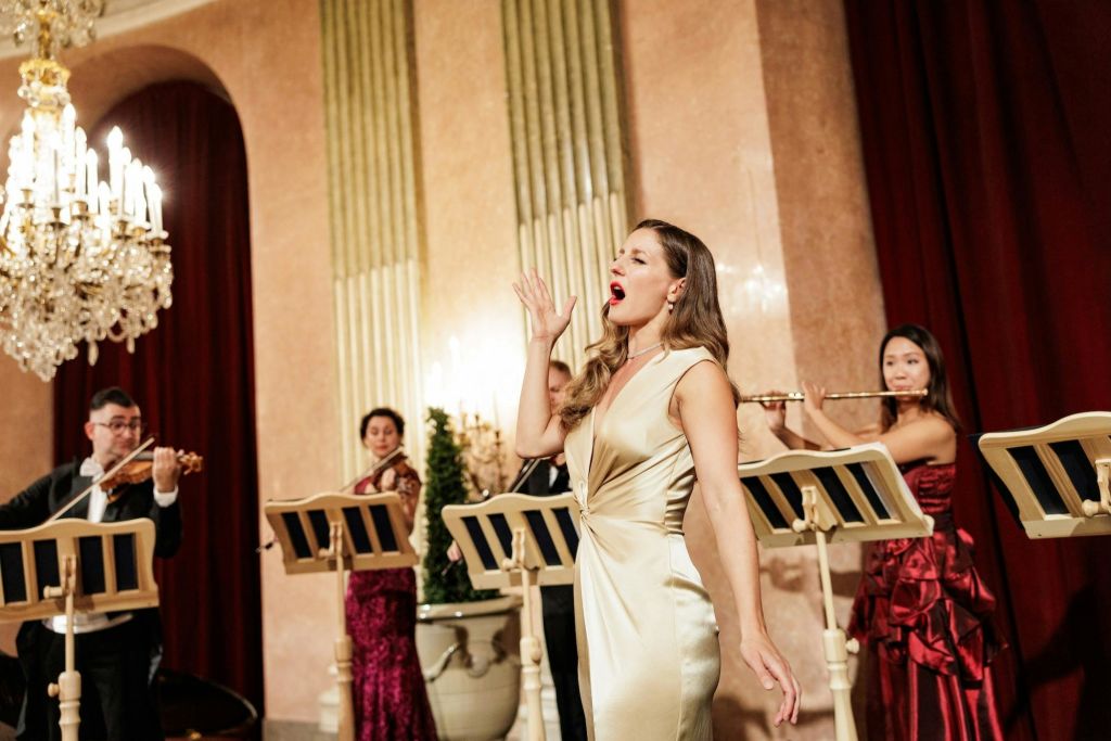 우아한 드레스를 입은 여성이 무대에서 열정적으로 노래하고 음악가와 함께 화려한 홀에 서 있습니다.