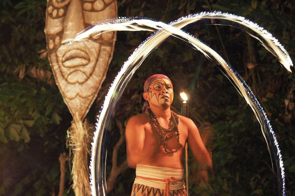 얼굴 페인트와 전통 복장을 한 사람이 부족 가면을 배경으로 밤에 불춤을 추고 있습니다.