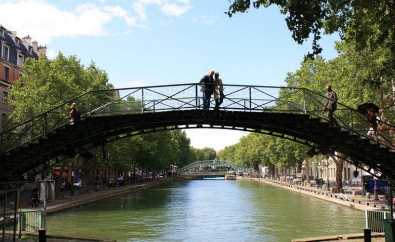 나무가 늘어선 운하를 배경으로 아치형 금속 다리에 서 있는 두 사람.