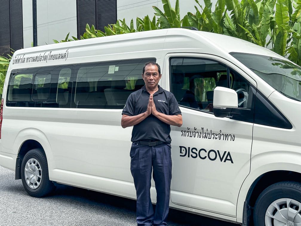 검은색 유니폼을 입은 한 남성이 "DISCOVA" 브랜드가 새겨진 흰색 밴 앞에서 정중한 제스처로 손을 맞잡고 서 있습니다.