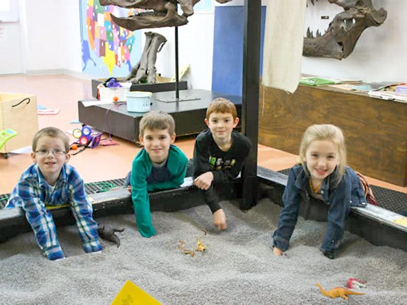 박물관 전시관에서 장난감 공룡과 함께 모래밭에서 놀고 있는 네 명의 어린이.