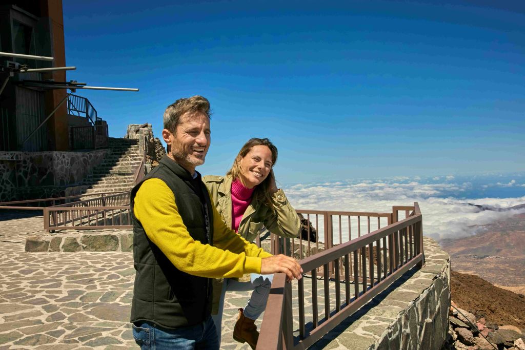 돌로 된 통로와 계단이 있는 전망대 난간에 기대어 웃고 있는 두 사람. 아래는 구름, 위는 푸른 하늘.