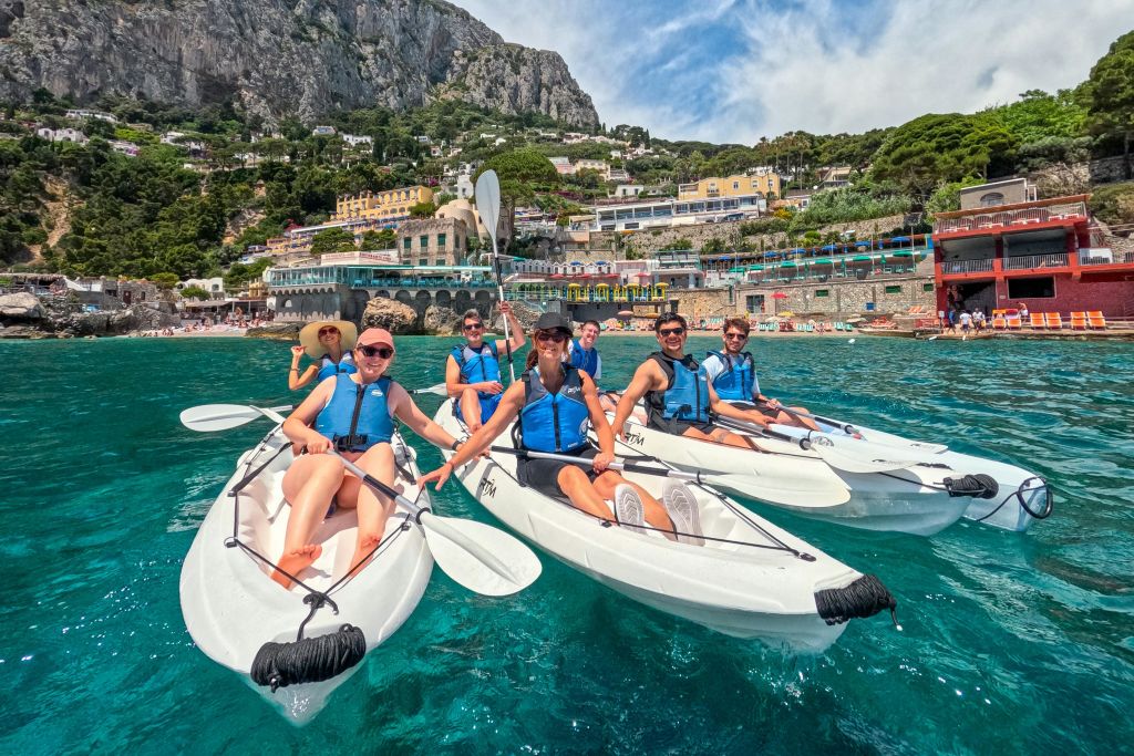푸른색 구명 조끼를 입은 한 무리의 사람들이 깨끗한 바다에서 카약을 타며 화려한 건물과 절벽을 배경으로 노를 젓고 있습니다.