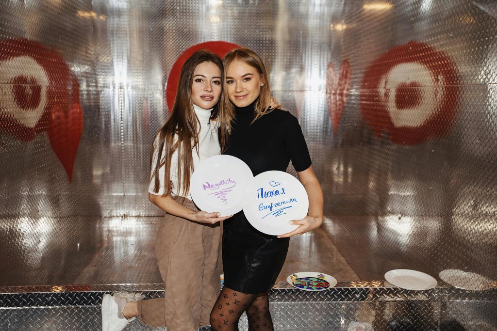 두 명의 청녀가 손으로 쓴 메시지가 담긴 흰색 접시를 들고 함께 포즈를 취하고 있다. 배경에는 하트 디자인의 금속 벽이 있습니다.