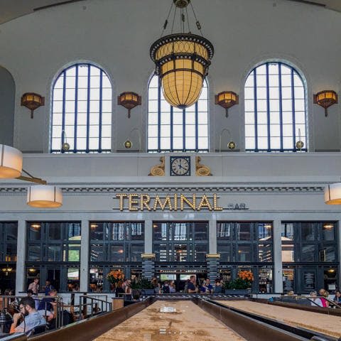 '터미널' 이라는 표지판이 있는 기차역 홀의 내부, 커다란 아치형 창문, 샹들리에, 사람들이 앉아 있습니다.