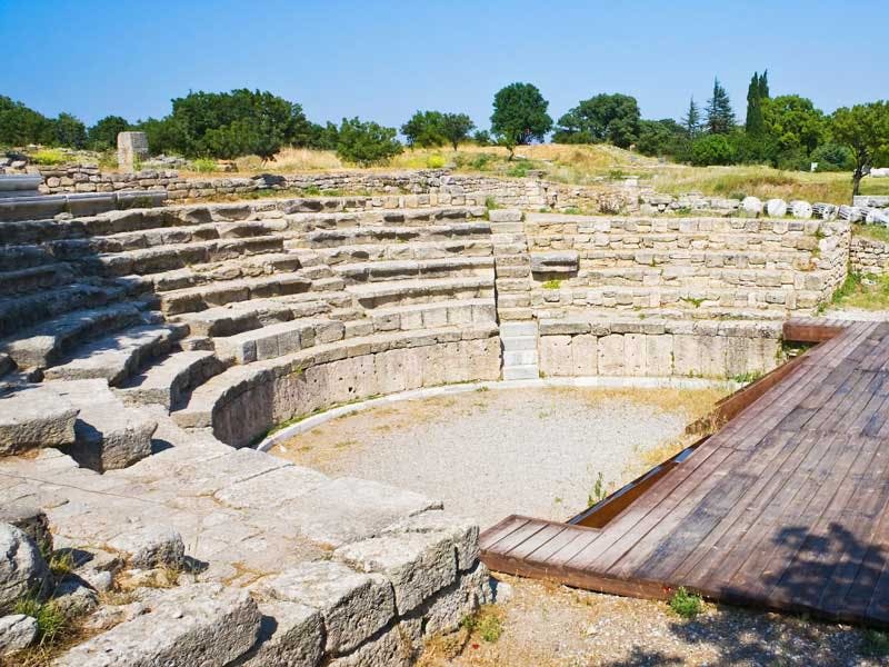 반원형 계단이 있는 고대 석조 원형극장은 잔디와 나무로 둘러싸여 있으며, 중심 지역으로 이어지는 나무 산책로가 있습니다.