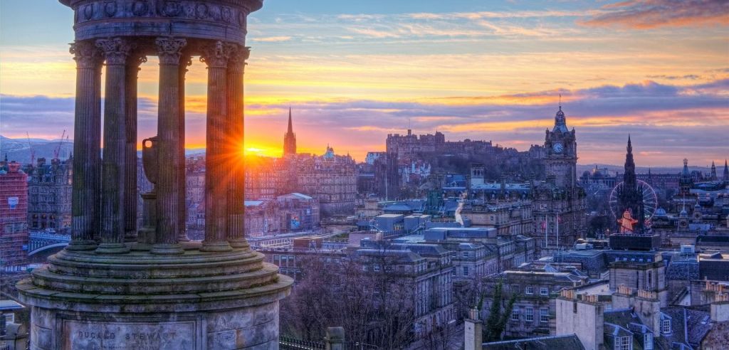 석양이 질 무렵 도심의 스카이라인이 한눈에 들어오며, 왼쪽에 돌기둥이 있는 유서 깊은 건물과 높은 첨탑이 보입니다.