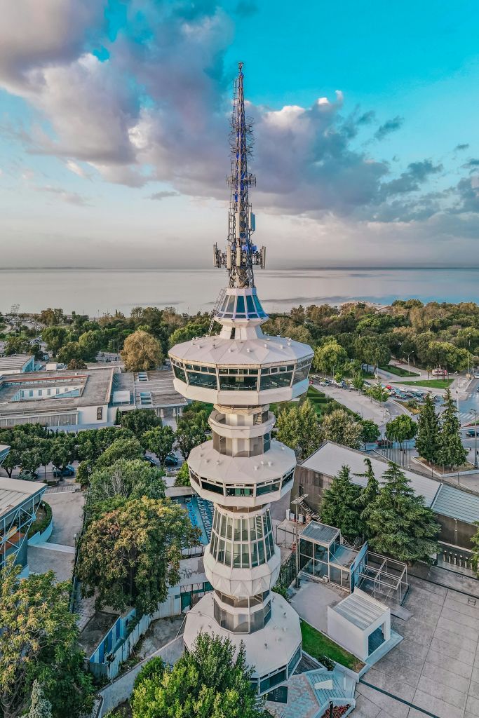 흐린 하늘 아래 물가 근처의 나무와 건물로 둘러싸인 원형 관측 플랫폼이 있는 높은 방송 타워.