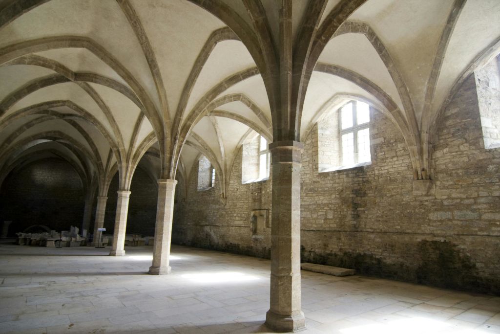 높은 아치형 천장과 여러 개의 기둥, 커다란 창문이 있는 석조 홀. 빛이 들어와 공간을 밝힙니다.