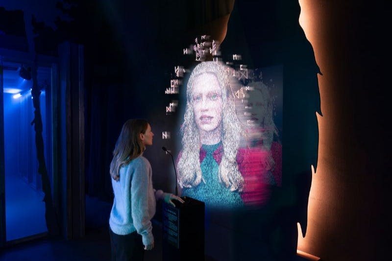 한 여성이 조명이 희미한 전시 공간에서 다른 사람의 홀로그램 투영과 상호 작용합니다.