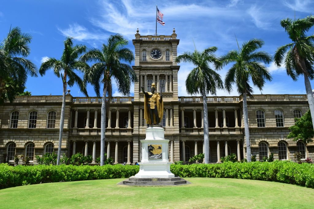중앙 시계탑과 미국 국기가 있는 화려한 역사적 건물입니다. 금색 가운을 입은 사람의 동상이 정면에 서 있습니다. 야자수가 줄지어 있습니다.