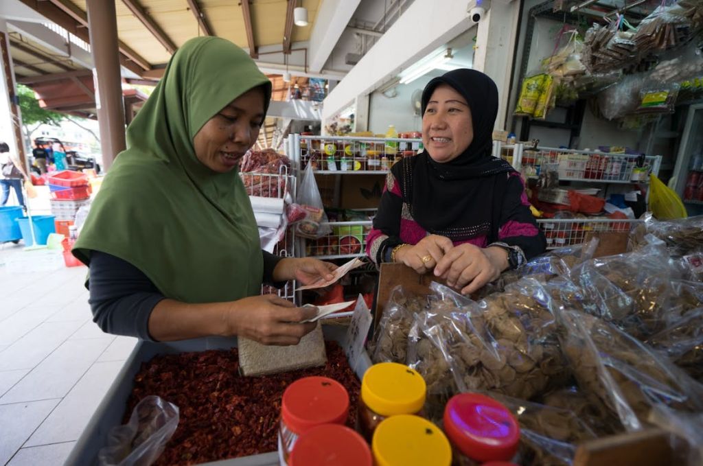 두 명의 여성이 다양한 향신료와 포장된 상품으로 가득한 시장 노점에서 상호 작용합니다. 한 여성이 작은 포장을 들고 있습니다.