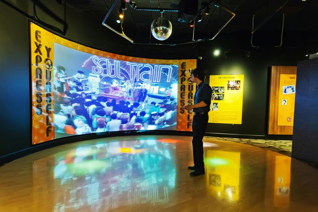 한 사람이 전시실에 서서 디스코 볼 아래에서 '소울 트레인' 동영상을 보여주는 대형 스크린을 보고 있습니다.