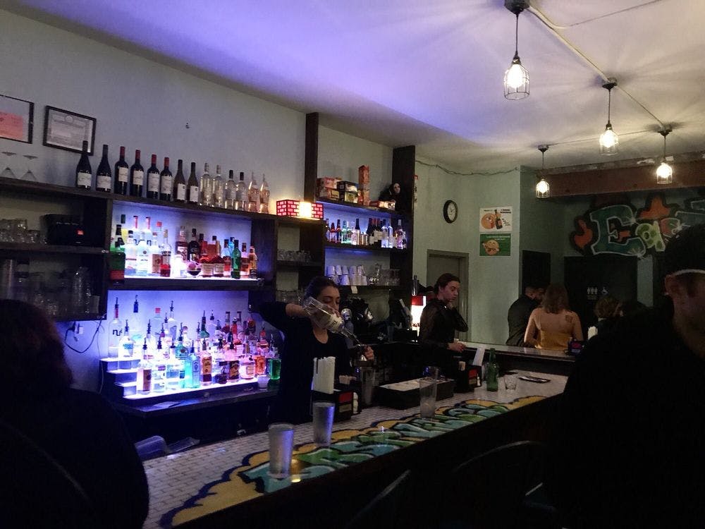 그래피티로 장식된 카운터가 있는 조명이 희미한 바, 바텐더가 음료를 붓고 있으며, 후원자들은 자리에 앉아 서 있습니다.