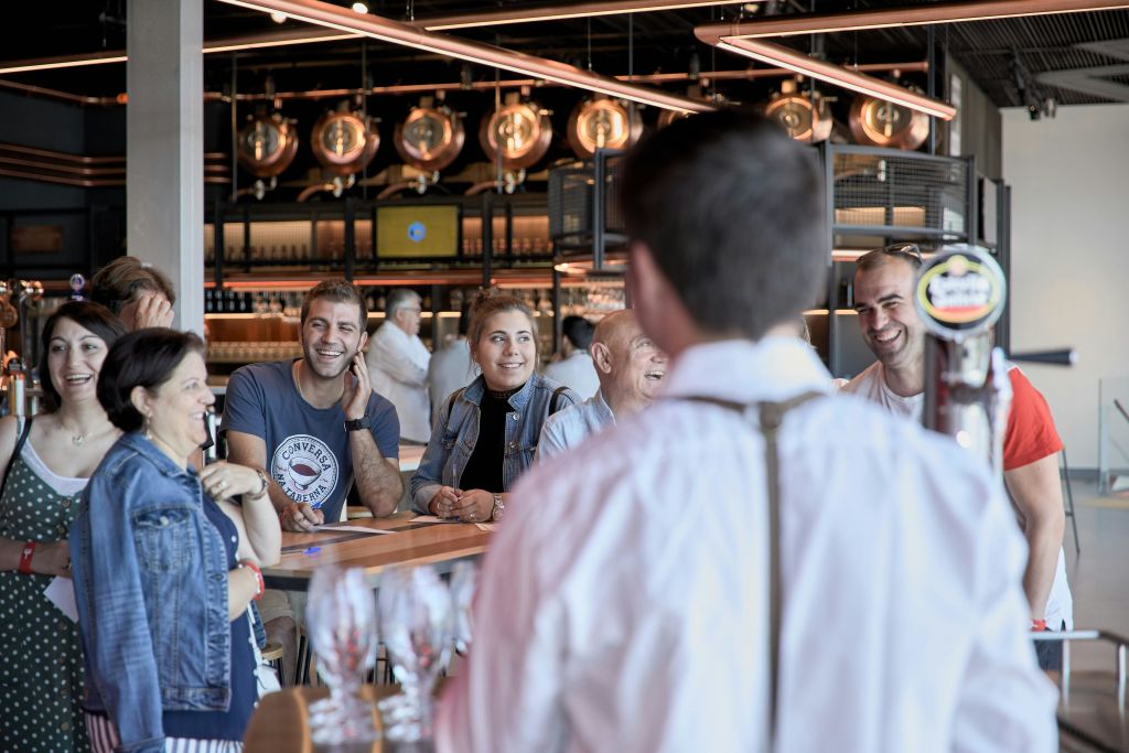 걸이식 조명과 구리 양조 탱크가 있는 현대적인 바에서 한 무리의 사람들이 나무 테이블을 둘러보며 미소를 짓고 대화를 나누고 있습니다.