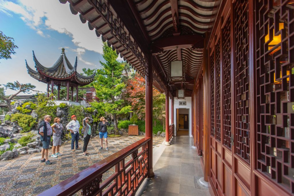 타일로 마감된 정자, 활기찬 나무, 붉은 나무 복도가 있는 중국 전통 정원에서 사진을 찍는 사람들.