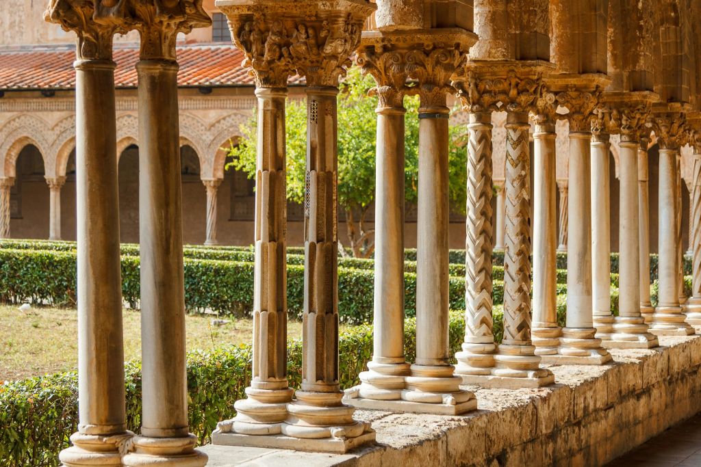 깔끔하게 다듬어진 울타리와 나무가 있는 울창한 정원이 내려다보이는 복도에 정교한 조각이 있는 화려한 석조 기둥이 있습니다.