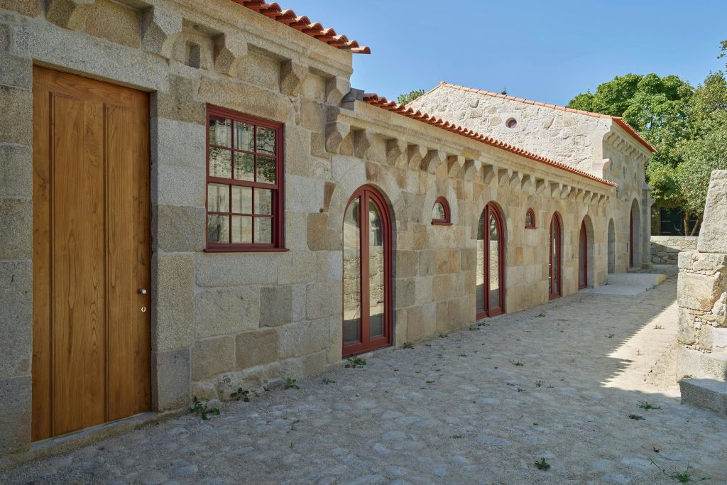 아치형 창문과 문, 빨간색 테두리, 타일 지붕이 있는 석조 건물. 앞쪽을 따라 자갈길이 이어집니다. 위쪽의 맑은 하늘.