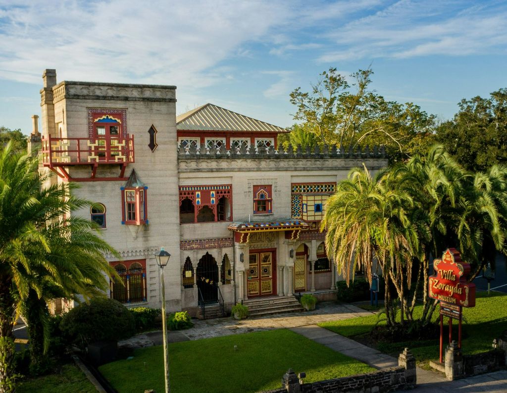 푸른 하늘 아래 야자수와 녹지로 둘러싸인 화려한 창문과 출입구가 있는 2층 건물입니다.