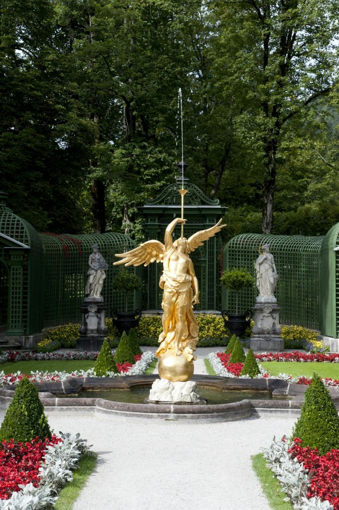 녹지와 장식 조각품으로 둘러싸인 조경된 정원에 창을 들고 있는 천사의 황금 동상이 서 있습니다.