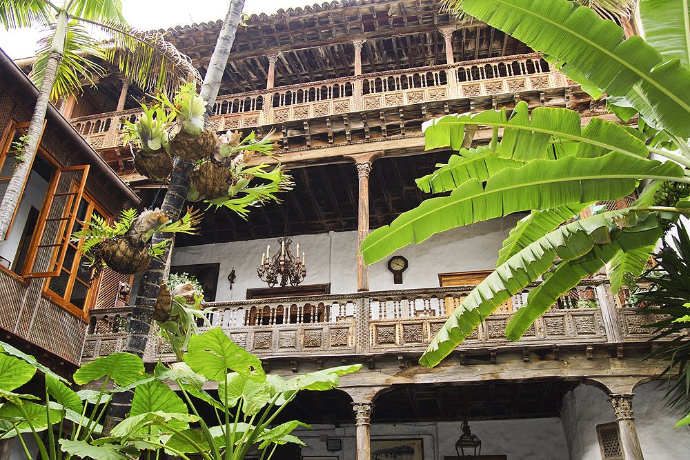 전면에 화려한 나무 발코니와 울창한 열대 식물이 있는 3층 건물. 2층에는 샹들리에가 걸려 있습니다.