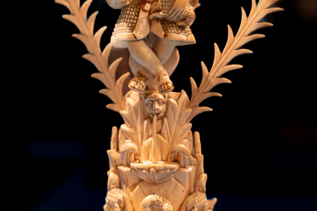 정교하게 조각된 아이보리 조각 위에 전통적인 복장을 한 인물이 있으며, 아래에는 디테일한 모티프와 얼굴이 있습니다.